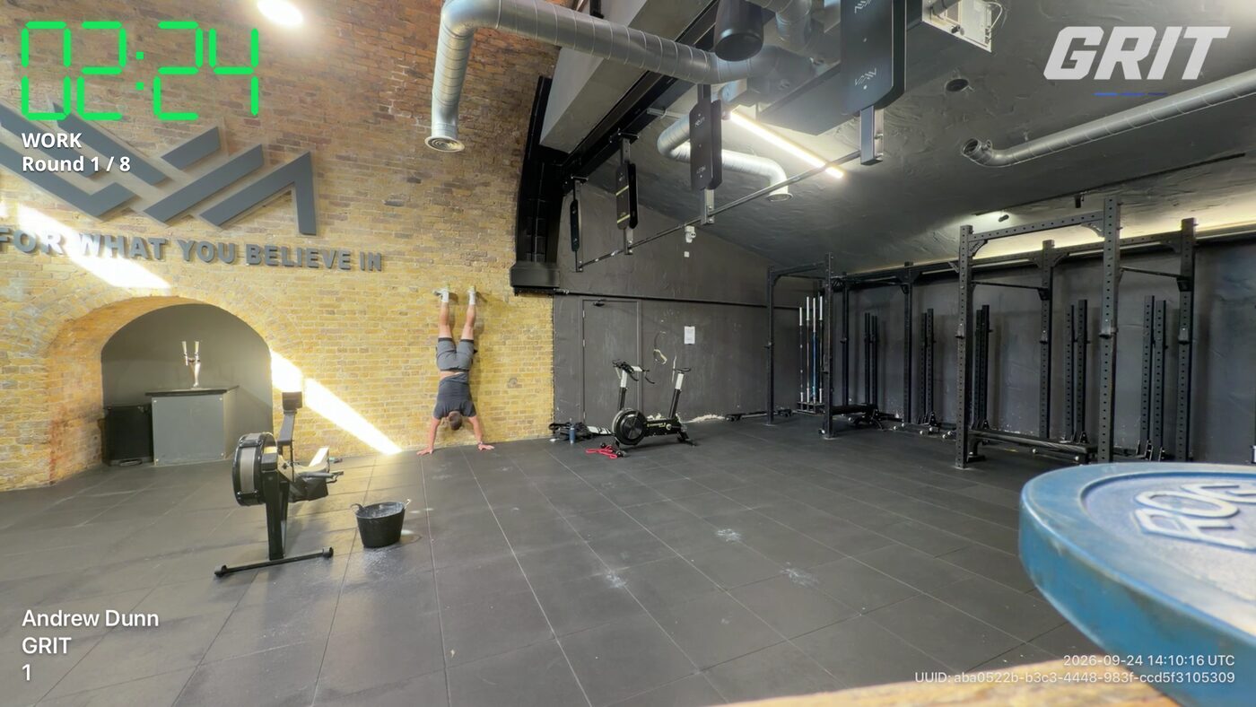A GRIT WOD Recorder frame from a gym session: an athlete doing a handstand hold, with the green clock at 02:24 and WORK Round 1 of 8 at the top left, the GRIT stamp at the top right, athlete and competition at the bottom left, and the date, time and video ID at the bottom right